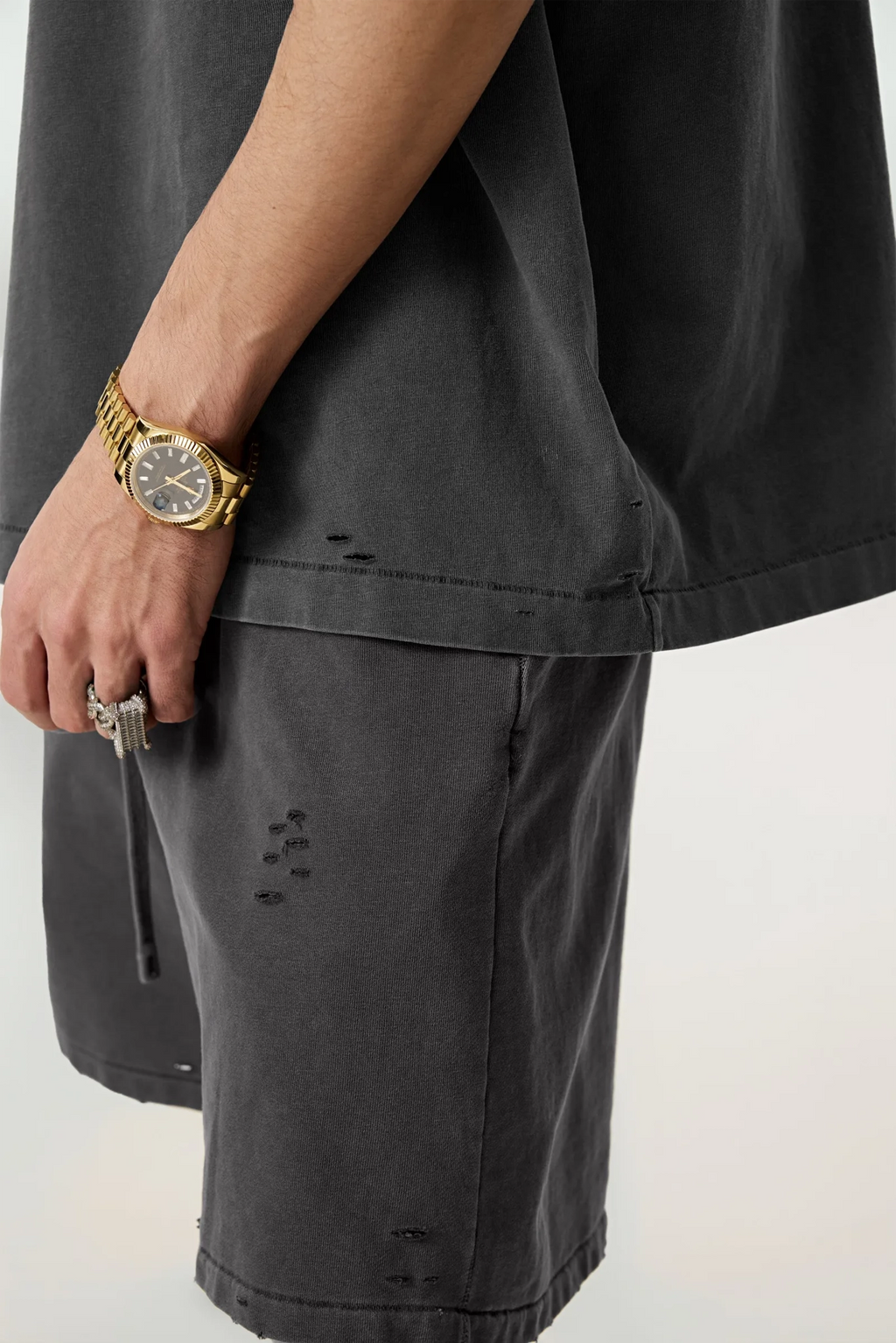Person wearing dark gray shorts with a gold watch and ring on a light background