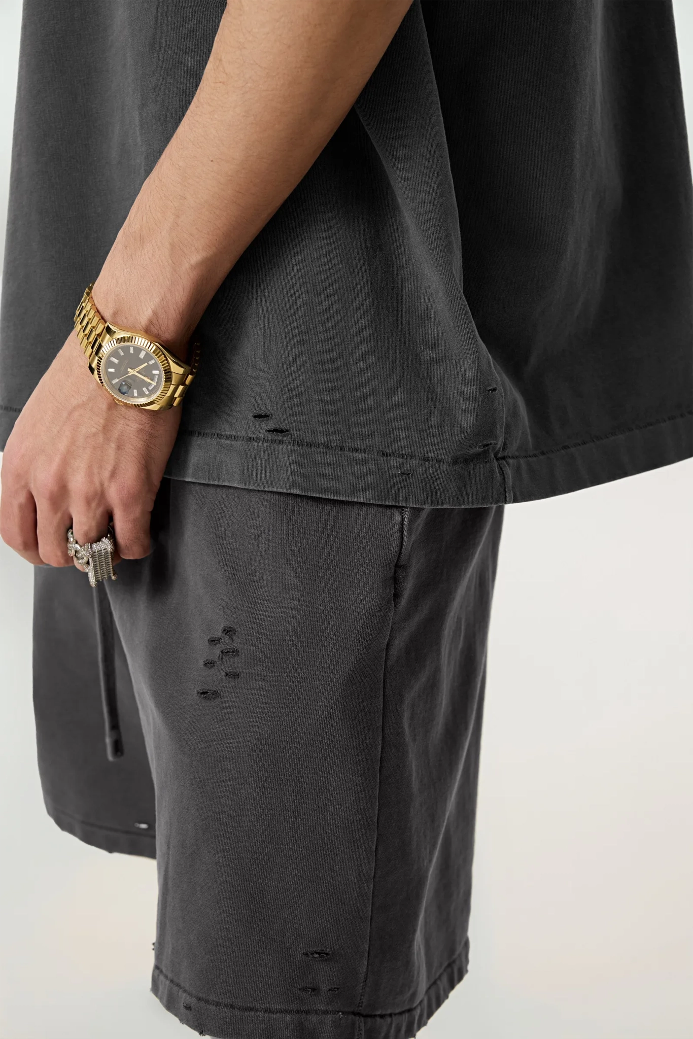 Person wearing dark gray shorts with a gold watch and ring on a light background