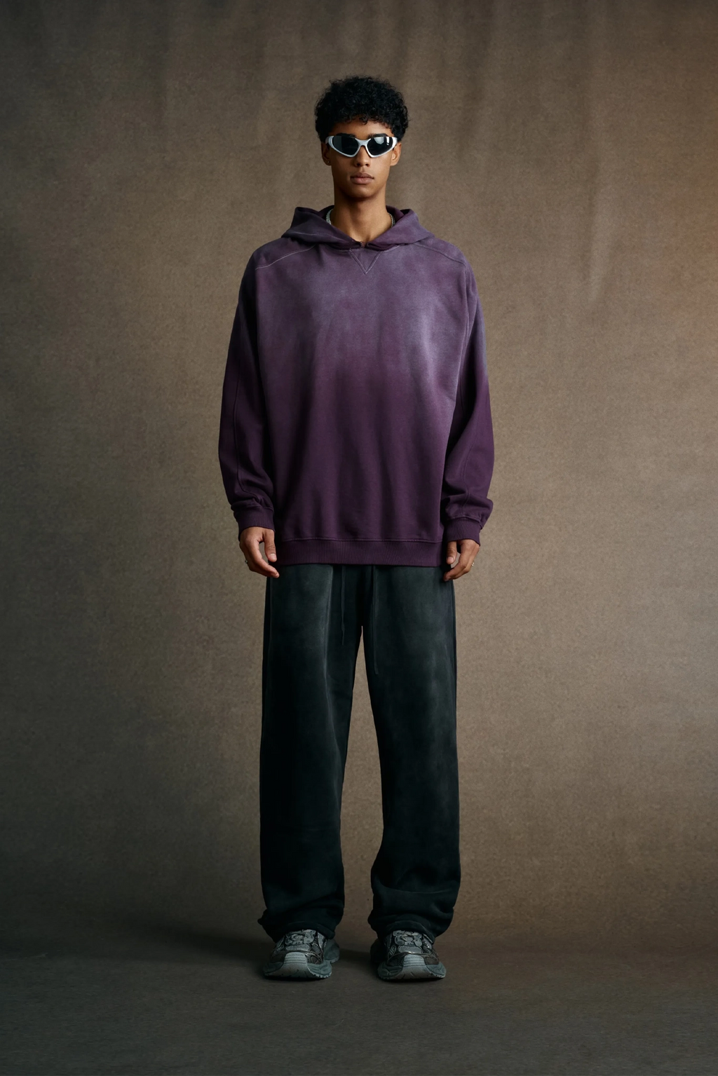 Person wearing a purple hoodie and sunglasses against a brown background