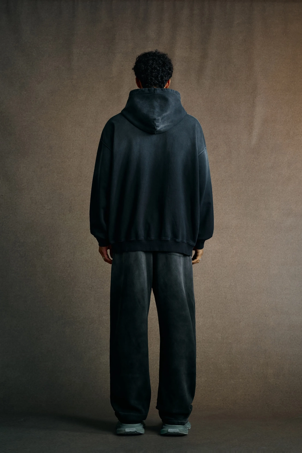 Person wearing a dark hoodie and pants standing against a plain background