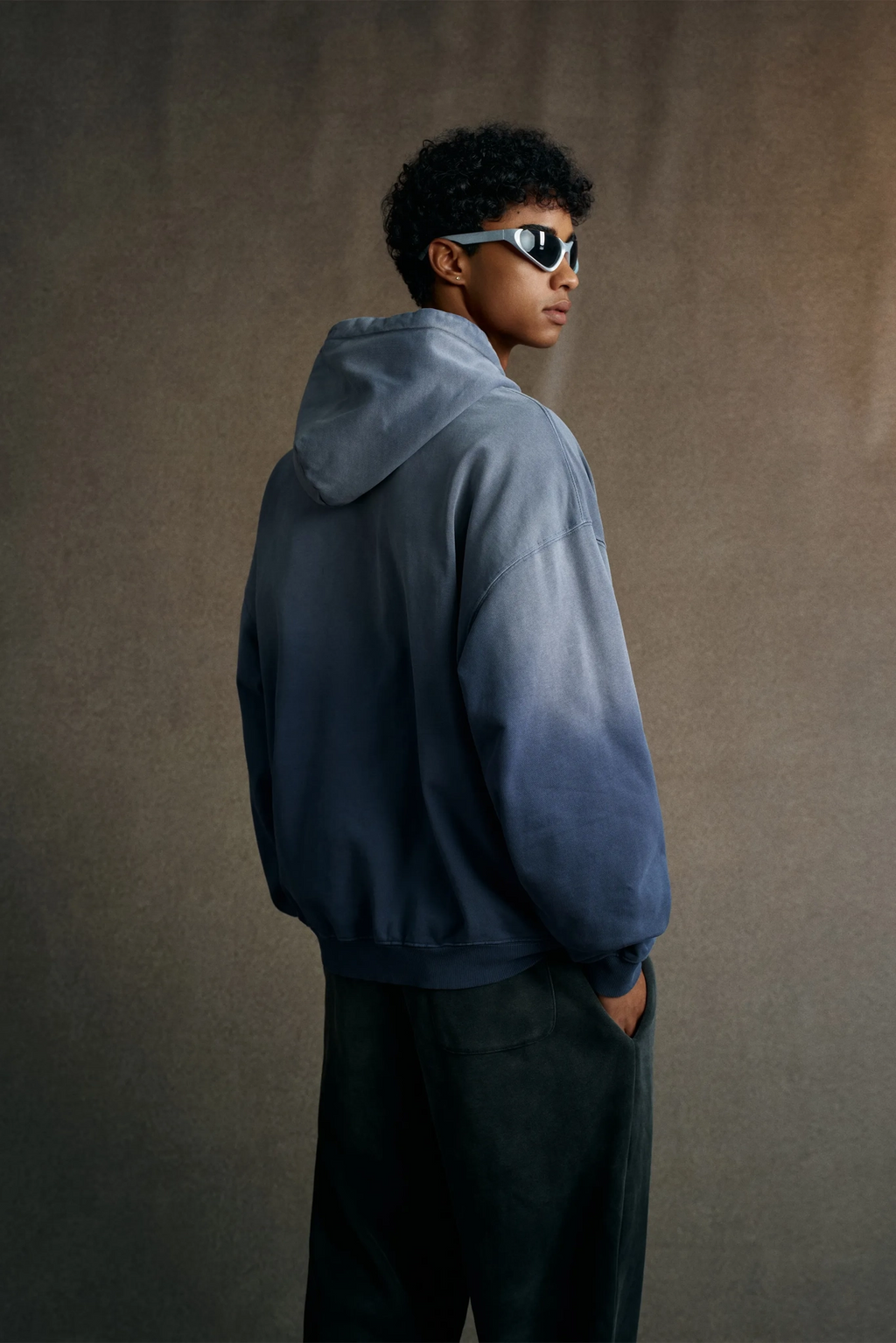 Person wearing a blue hoodie and sunglasses against a brown background