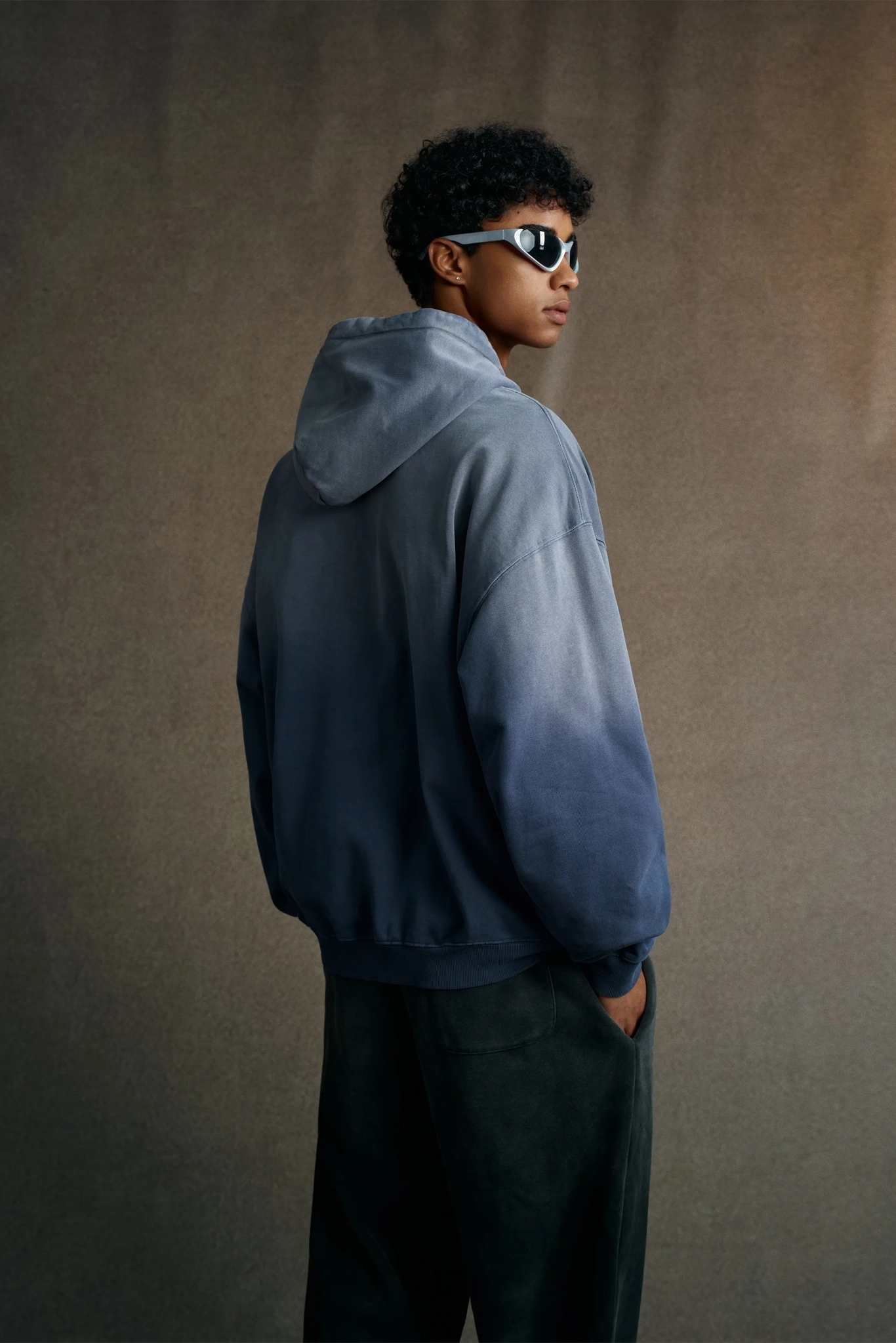 Person wearing a blue hoodie and sunglasses against a brown background