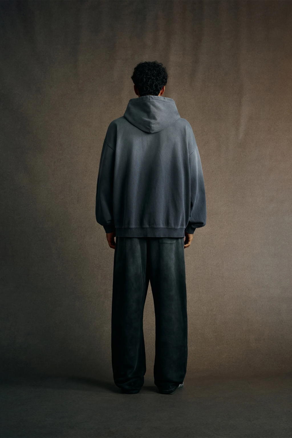 Person wearing a gray hoodie and dark pants standing against a plain background