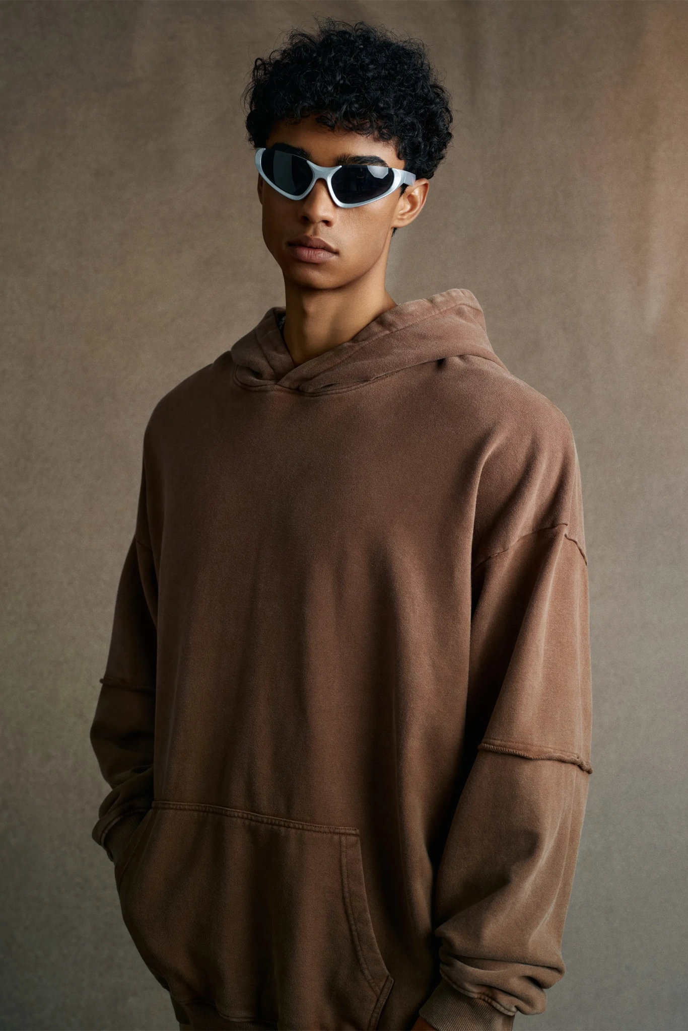 Person wearing a brown hoodie and sunglasses against a neutral background