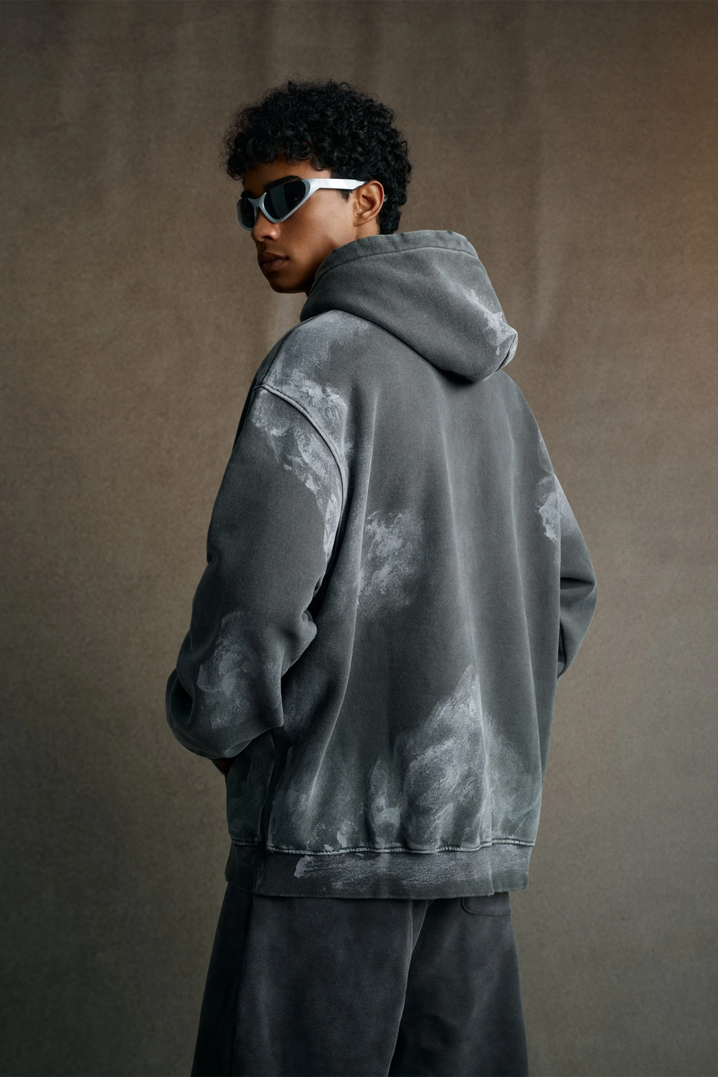 Person wearing a gray tie-dye hoodie and sunglasses on a brown background