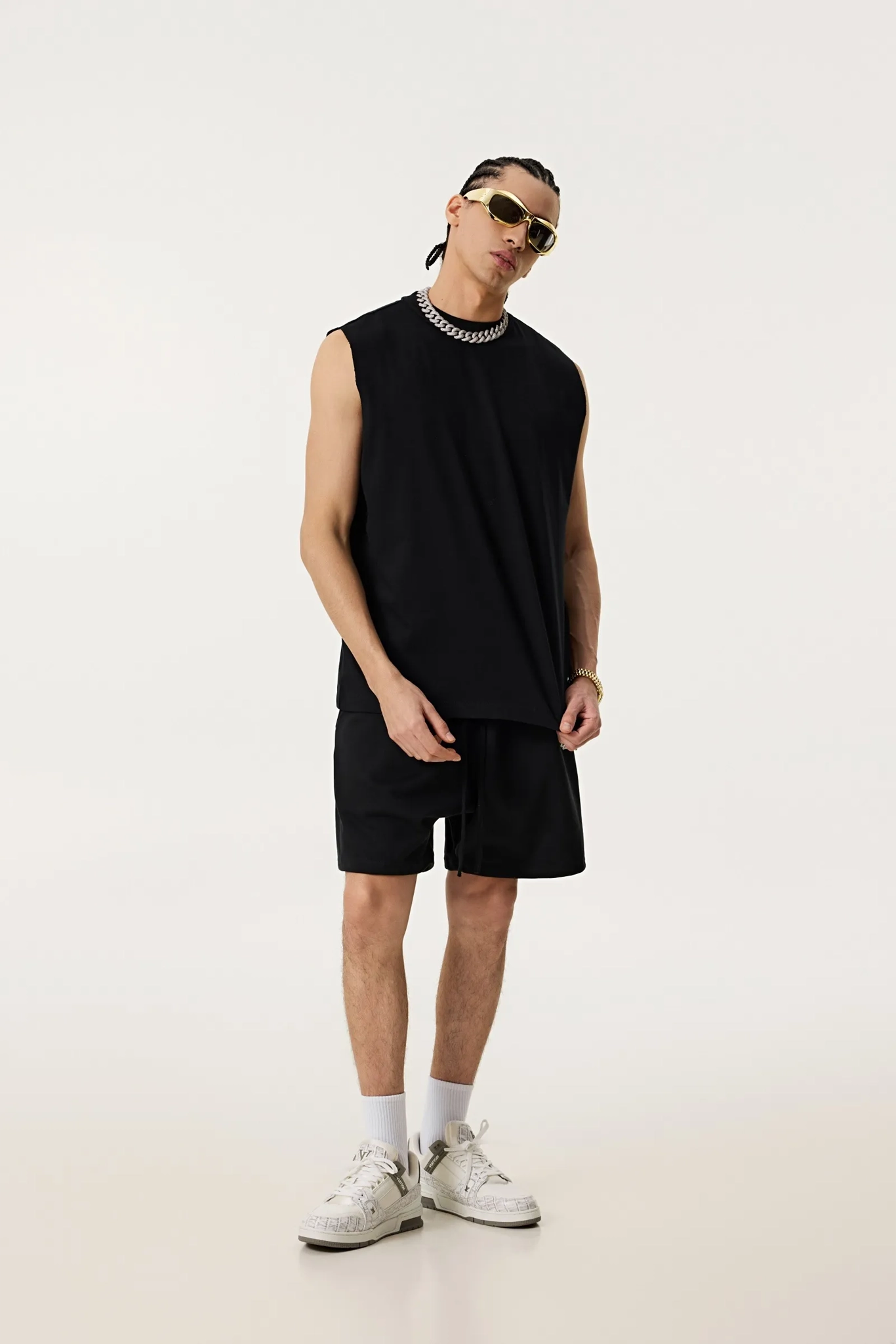 Person wearing a black sleeveless shirt, black shorts, white sneakers, and sunglasses on a white background