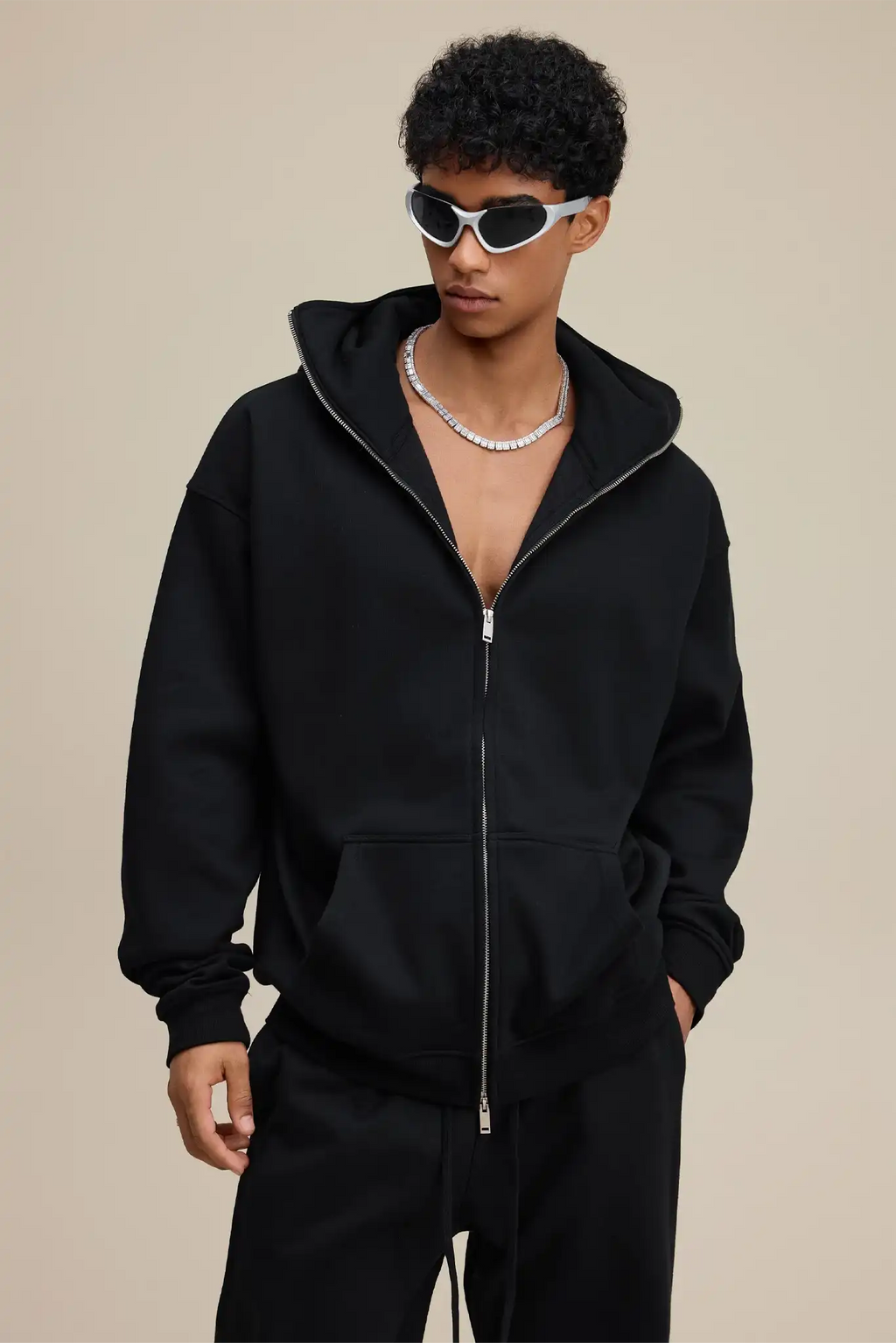 Person wearing a black hoodie and pants with sunglasses on a beige background