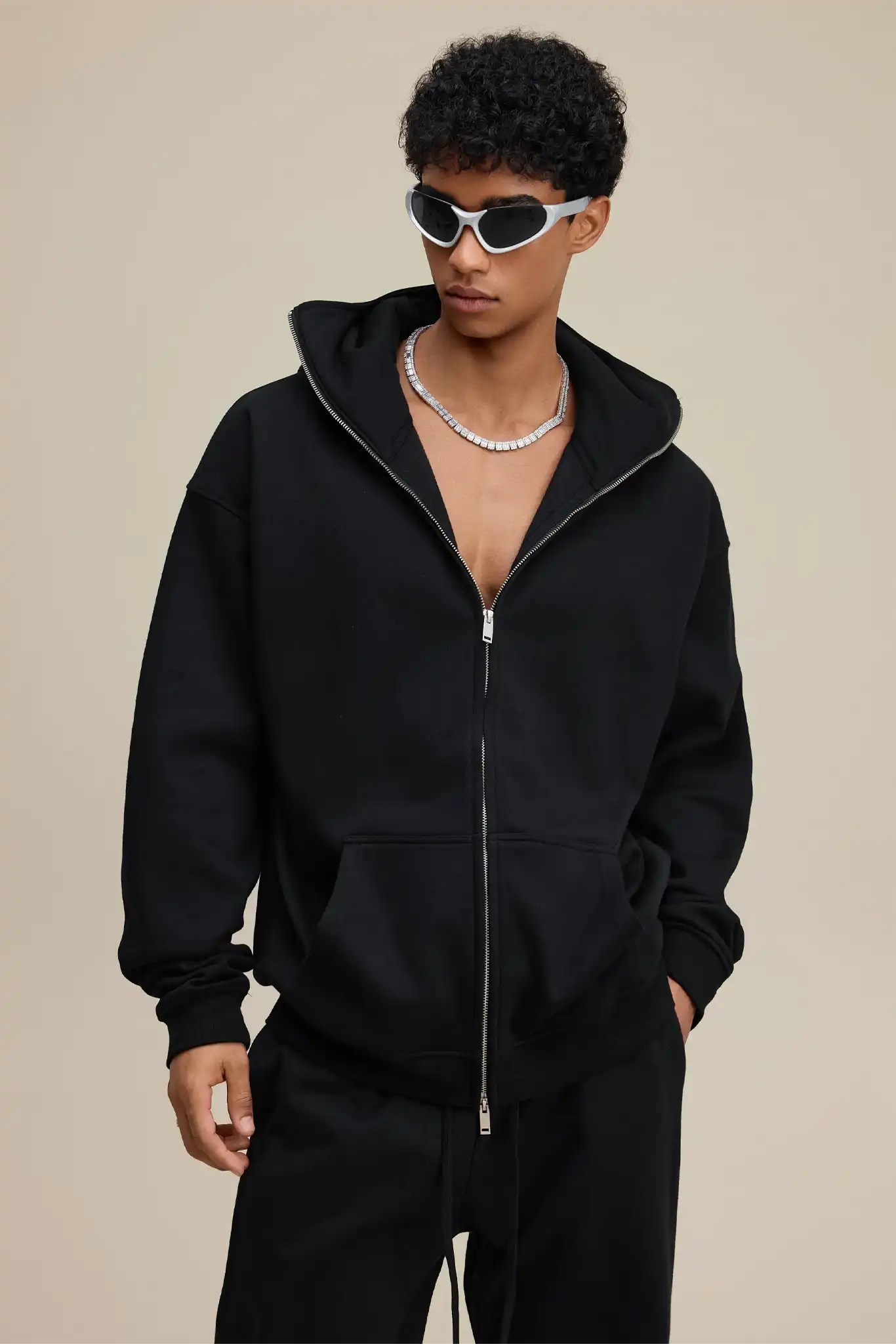 Person wearing a black hoodie and pants with sunglasses on a beige background