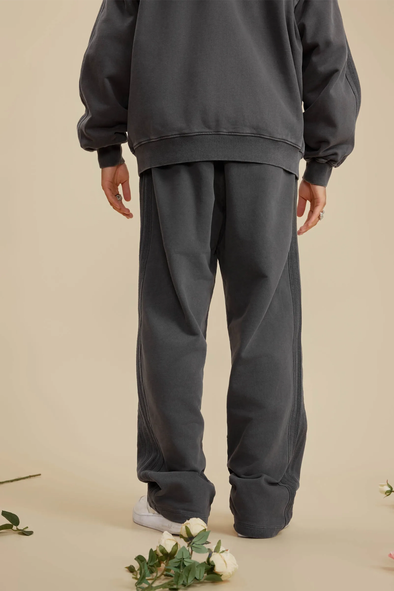 Person wearing a dark gray sweatshirt and pants on a beige background with flowers at the bottom.