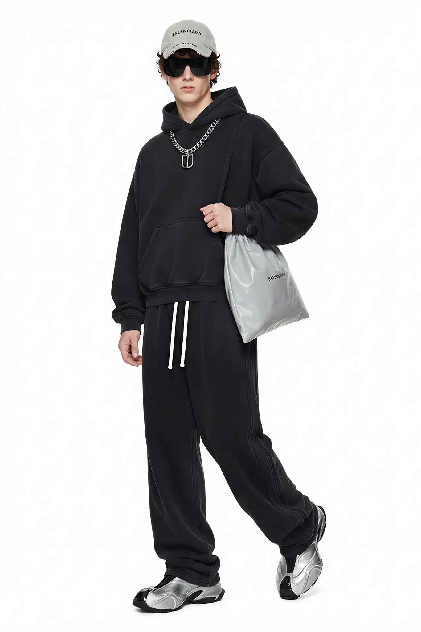 Person wearing a black hoodie and pants with a white cap and sunglasses, holding a gray bag on a white background.
