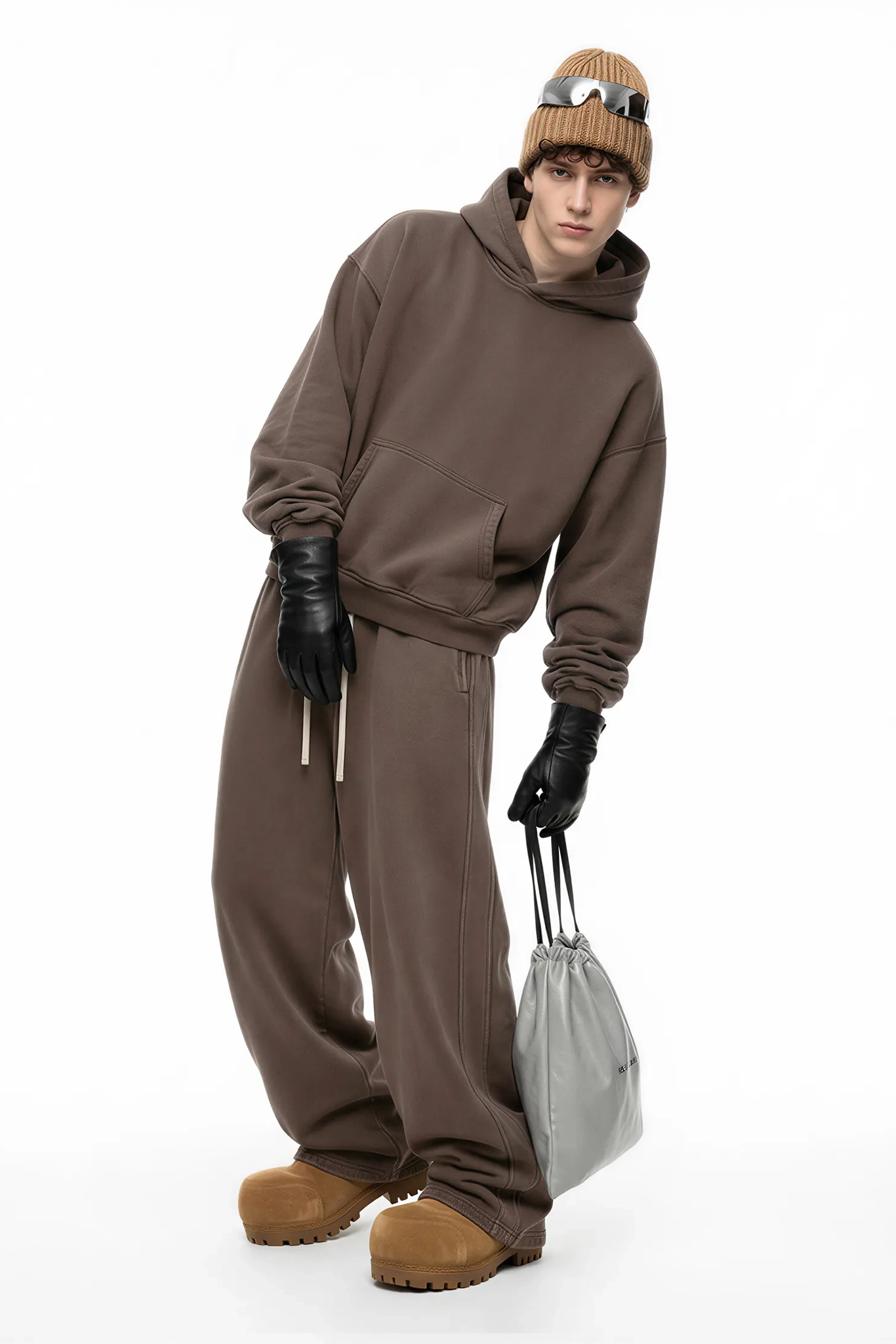 Person wearing a brown tracksuit with a beanie and gloves, holding a white bag on a white background
