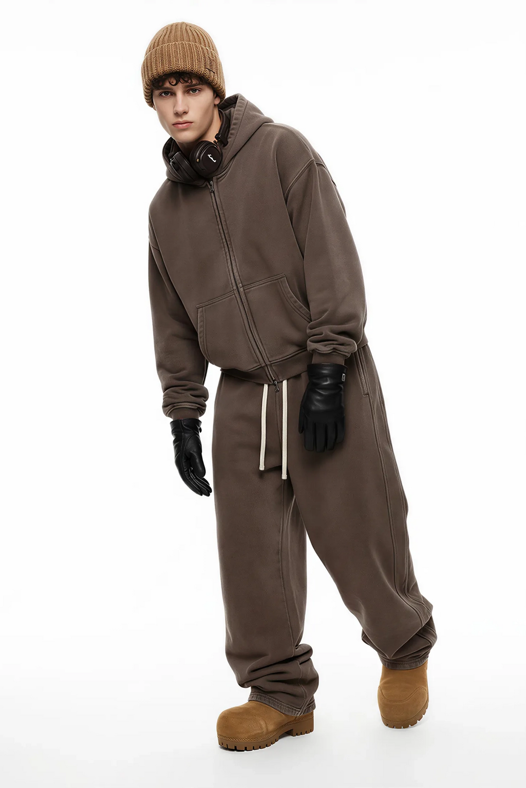 Person wearing a brown tracksuit with a beanie, headphones, and boots on a white background