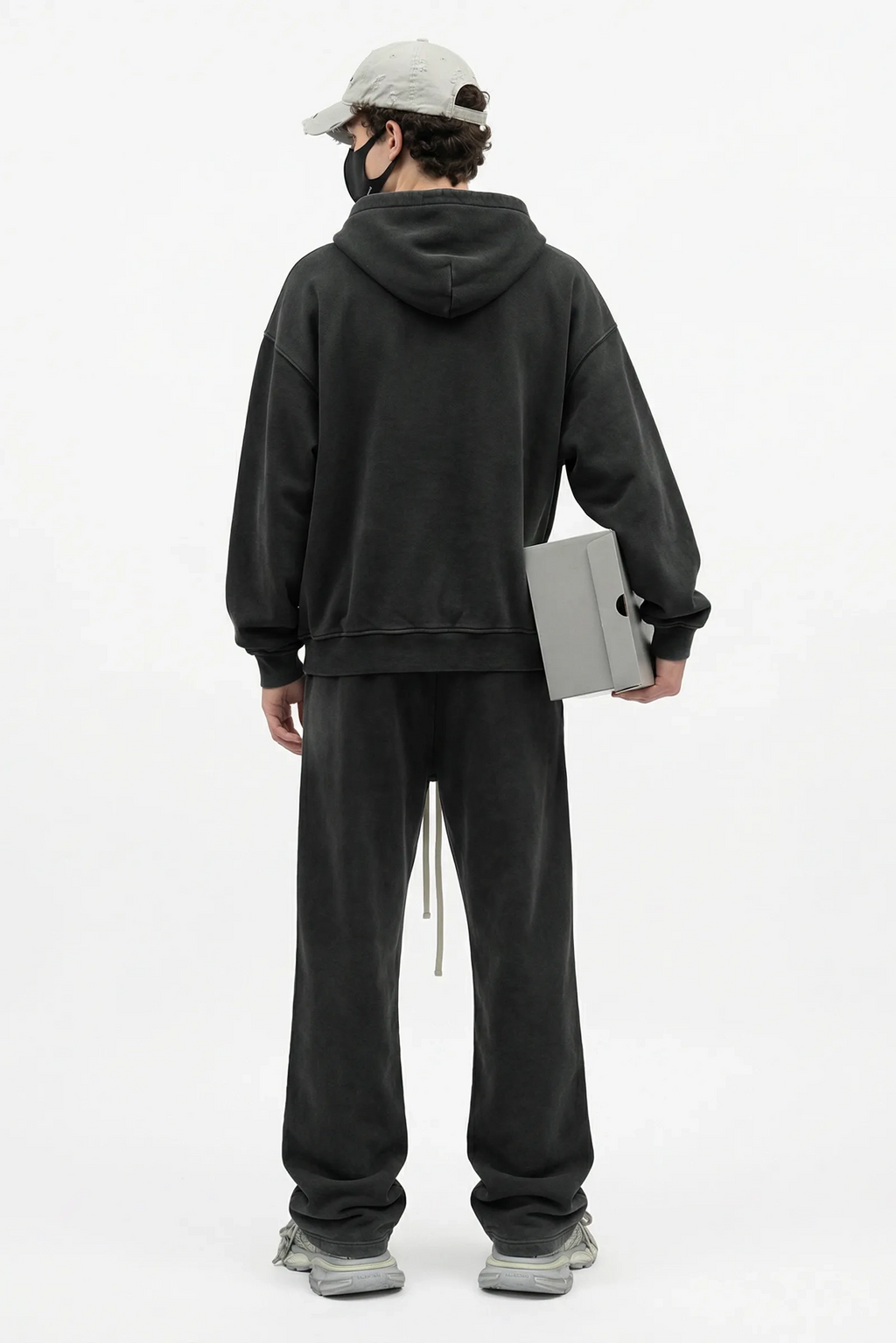 Person wearing a black hoodie and pants holding a gray box on a white background