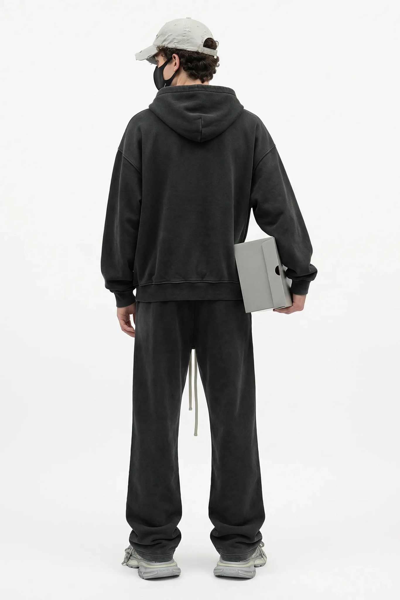 Person wearing a black hoodie and pants holding a gray box on a white background