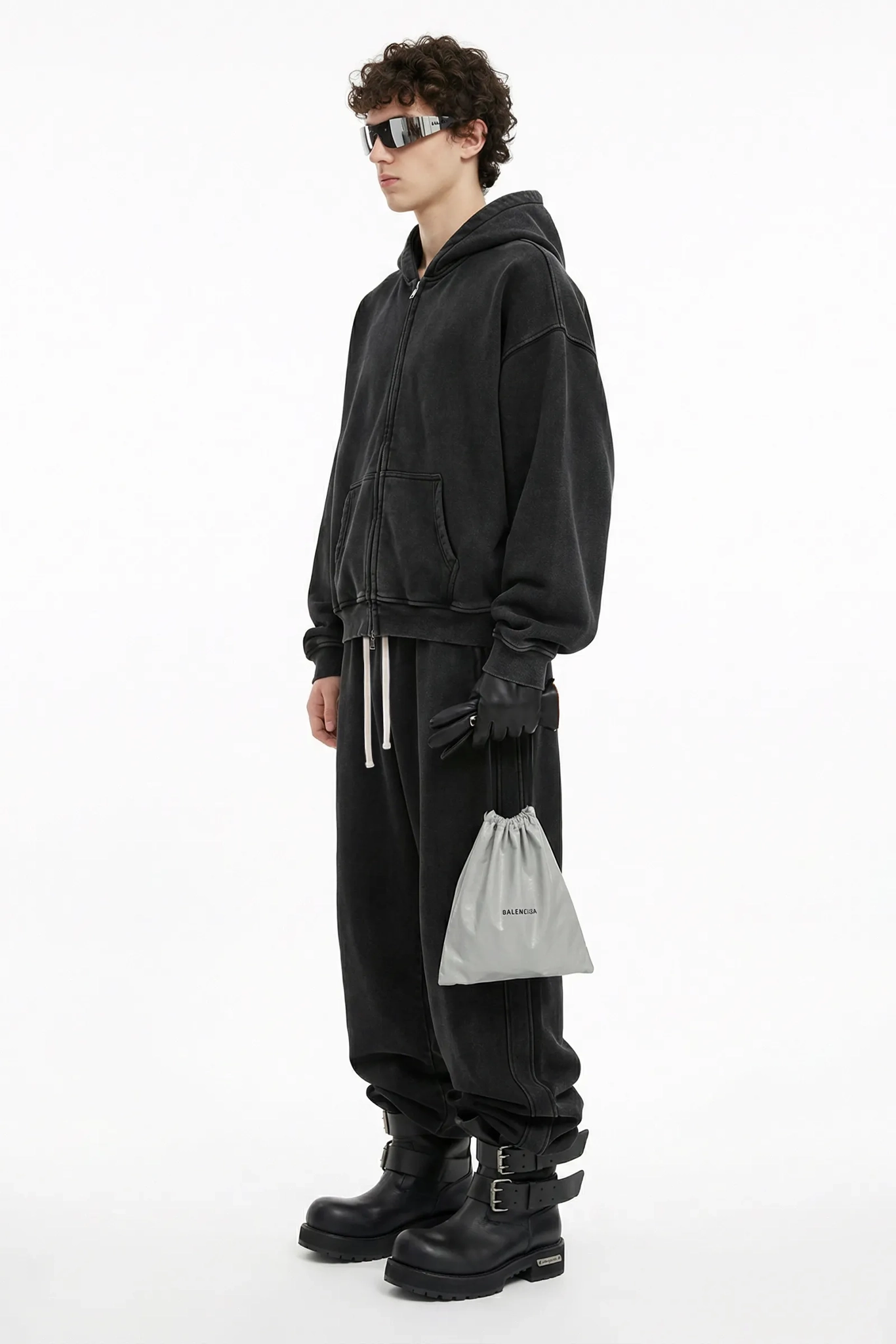 Person wearing a black outfit with sunglasses and holding a gray drawstring bag on a white background