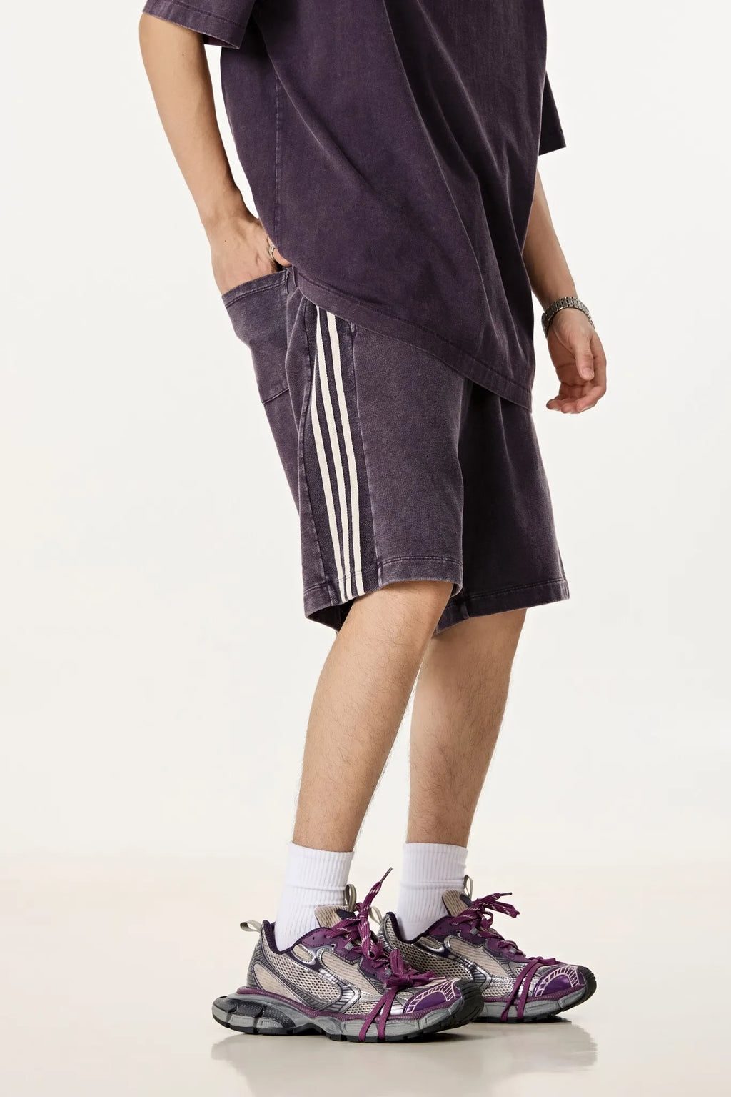 Person wearing a dark purple oversized t-shirt, shorts with white stripes, and purple sneakers on a white background