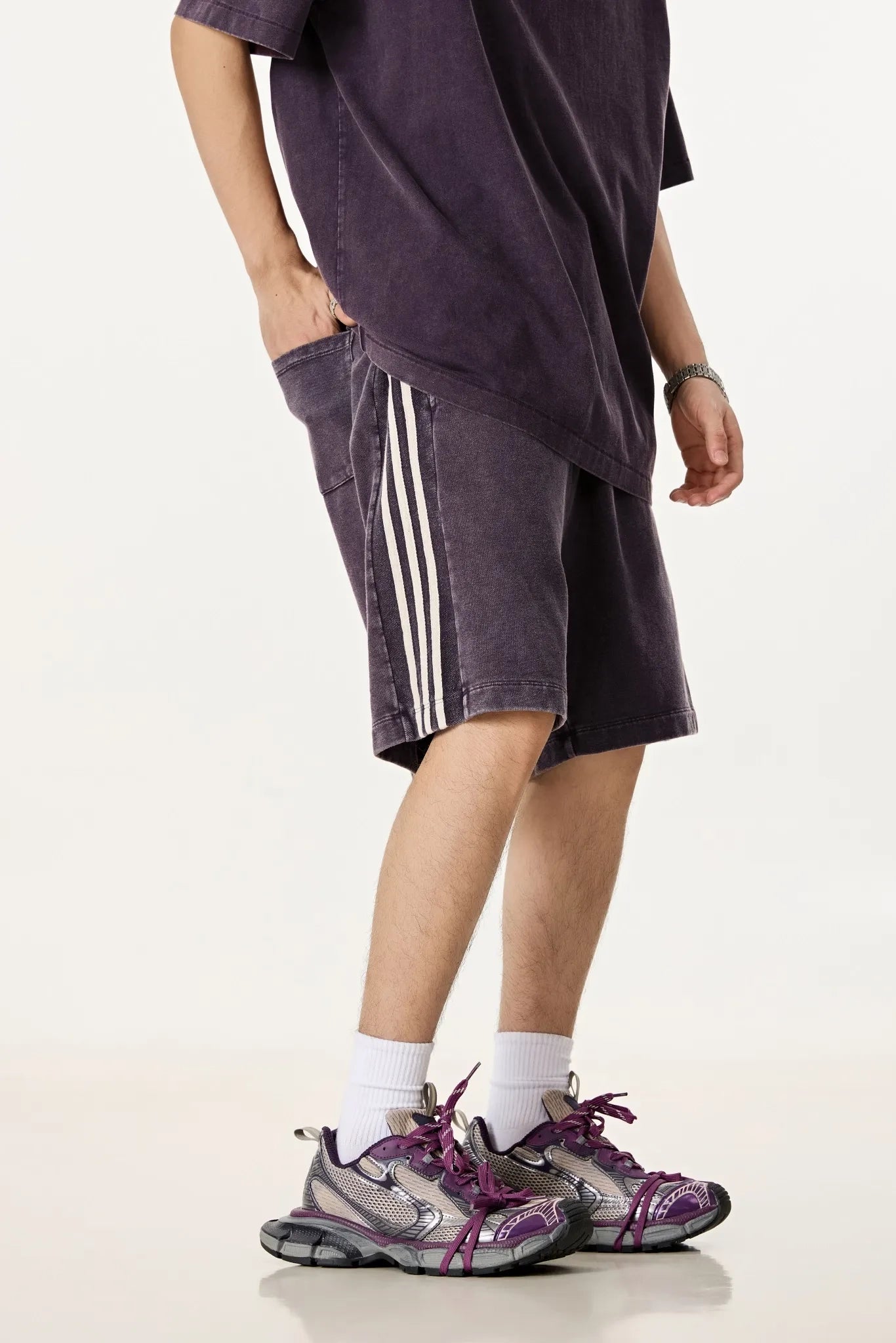 Person wearing a dark purple oversized t-shirt, shorts with white stripes, and purple sneakers on a white background