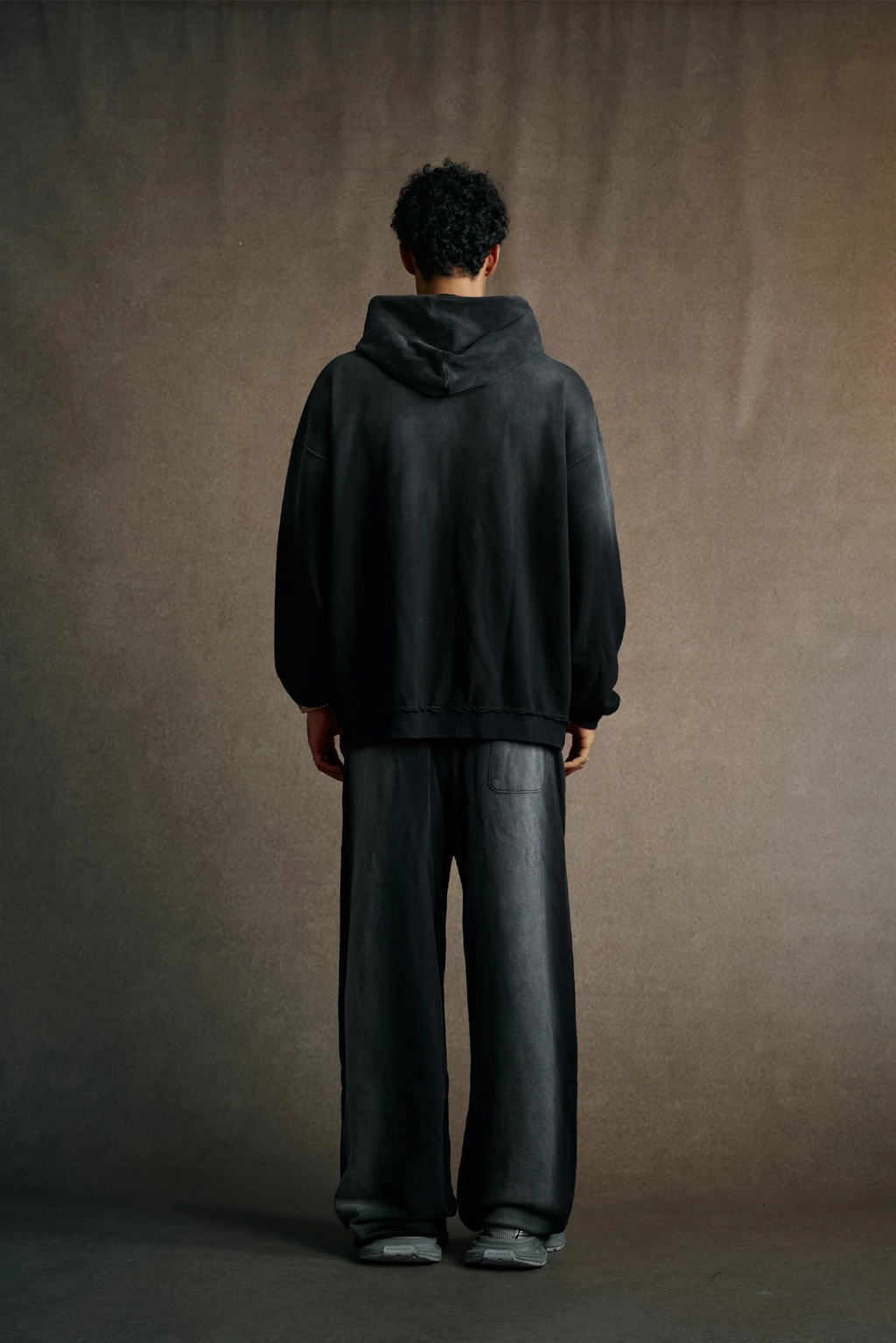 Person wearing a dark hoodie and pants standing against a plain background