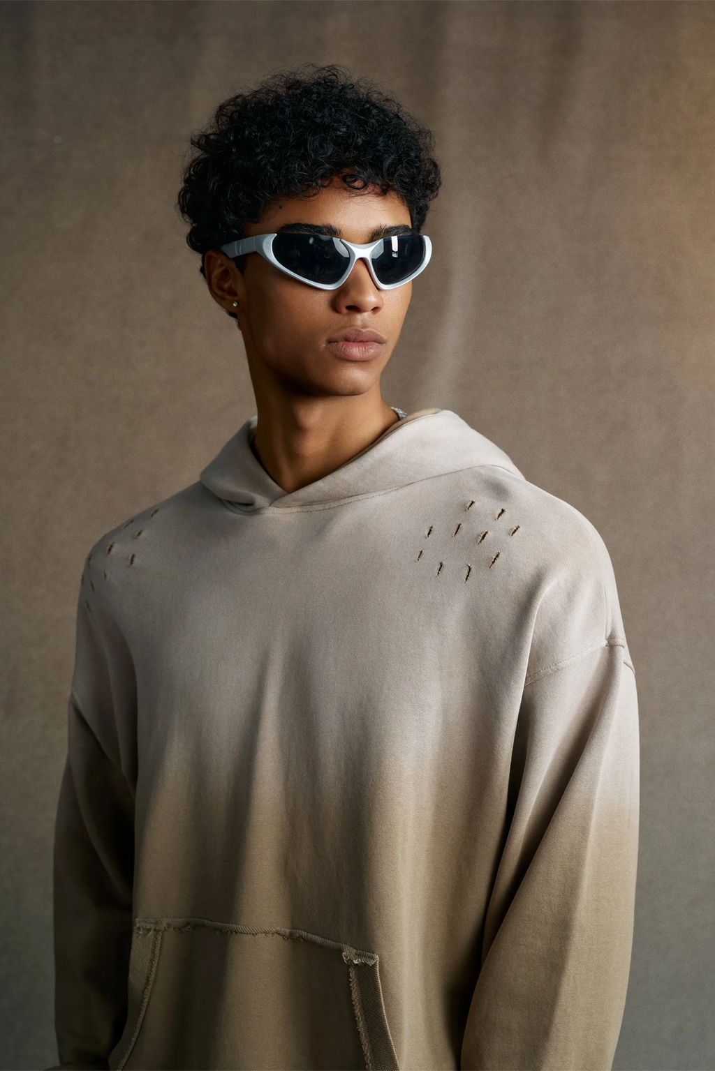 Person wearing a beige hoodie and sunglasses against a brown background