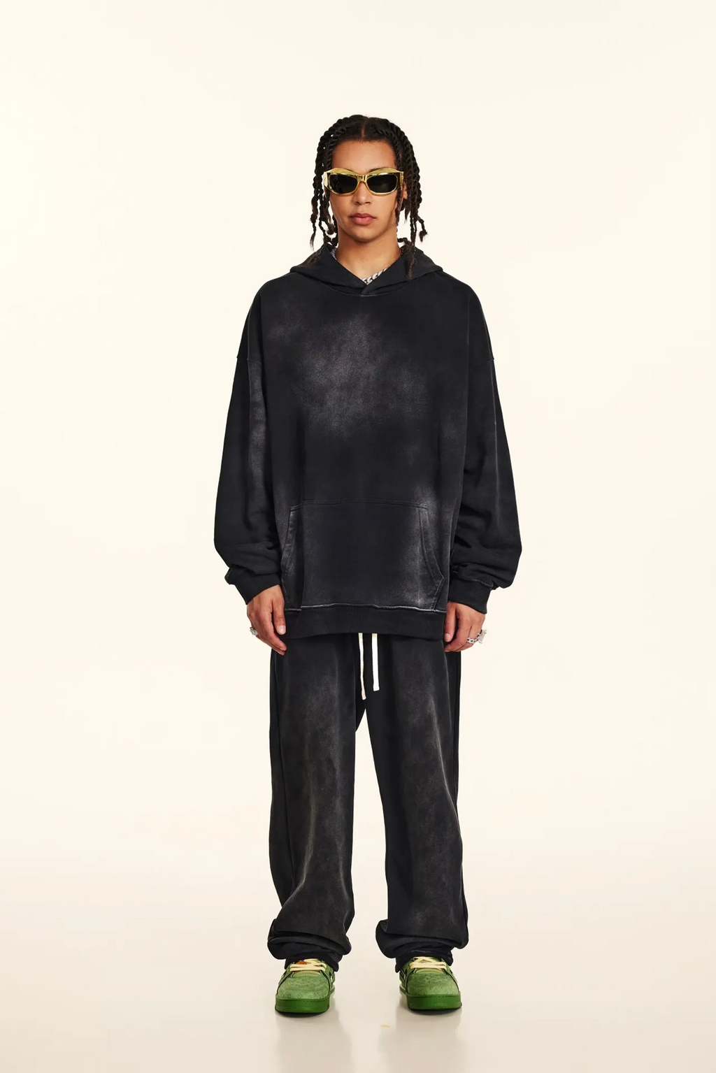 Person wearing a black hoodie and pants with green shoes on a white background