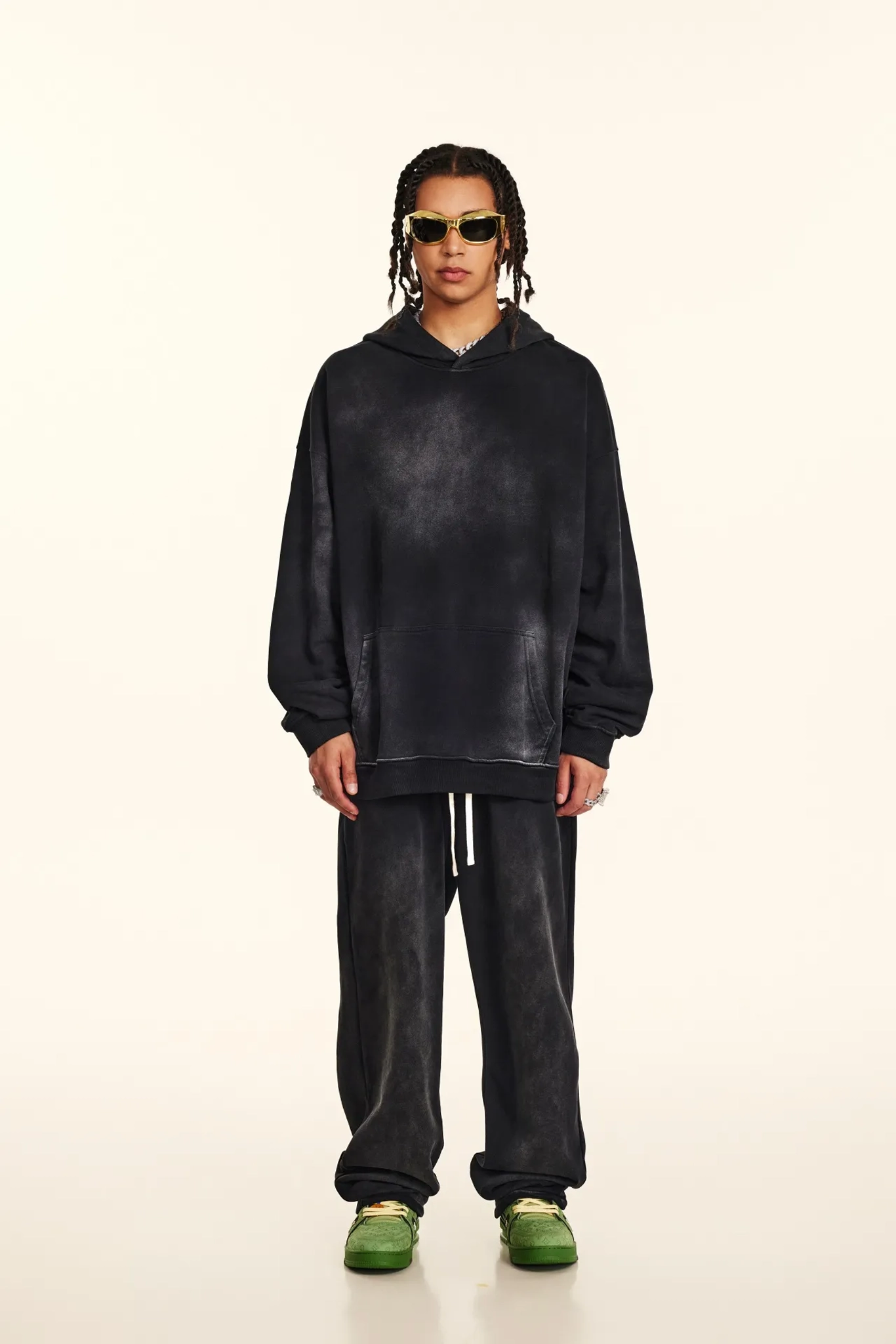 Person wearing a black hoodie and pants with green shoes on a white background