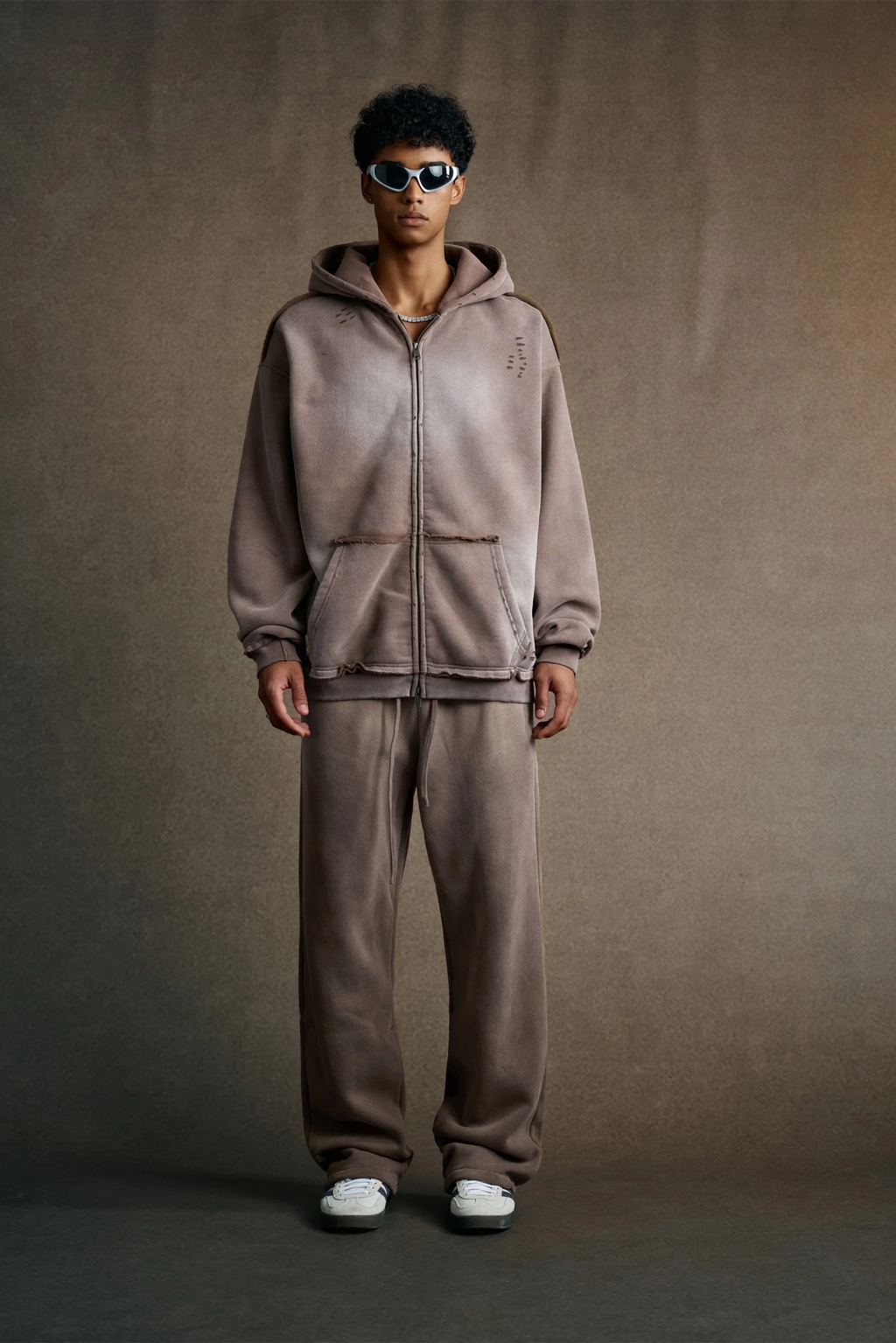 Man wearing a brown tracksuit and sunglasses against a plain background