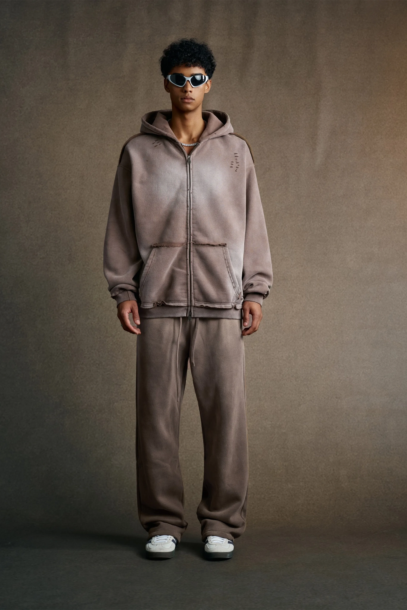 Man wearing a brown tracksuit and sunglasses against a plain background