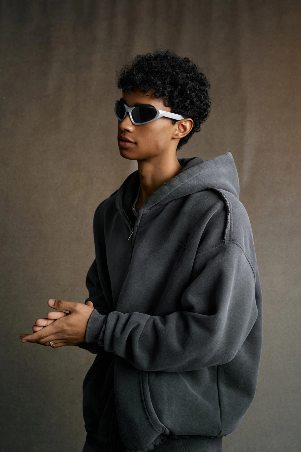 Person wearing a gray hoodie and sunglasses against a brown background