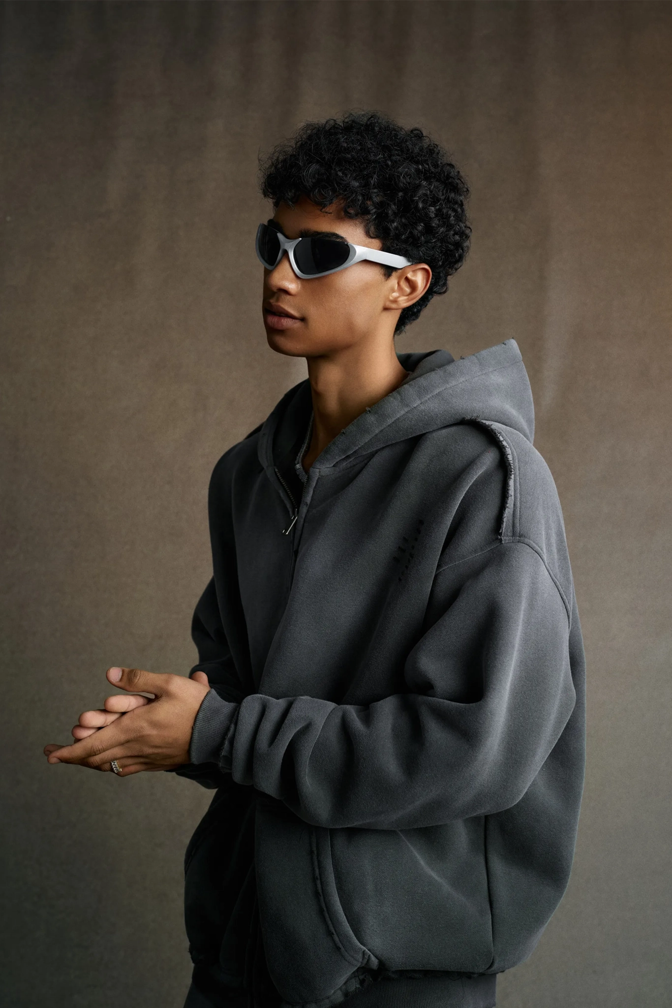Person wearing a gray hoodie and sunglasses against a brown background