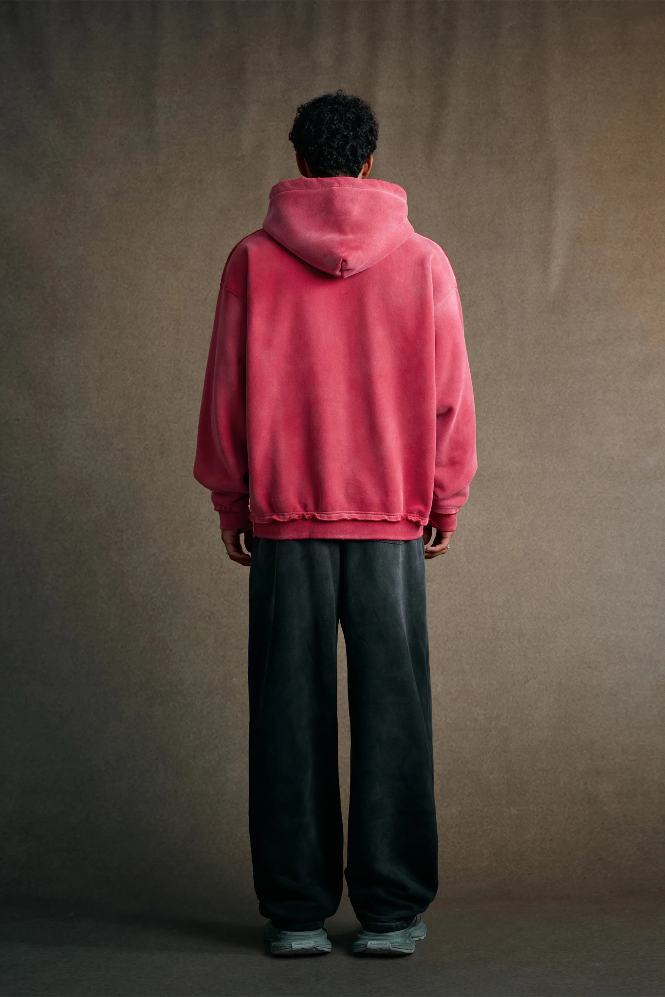 Person wearing a pink hoodie and dark pants standing against a brown background