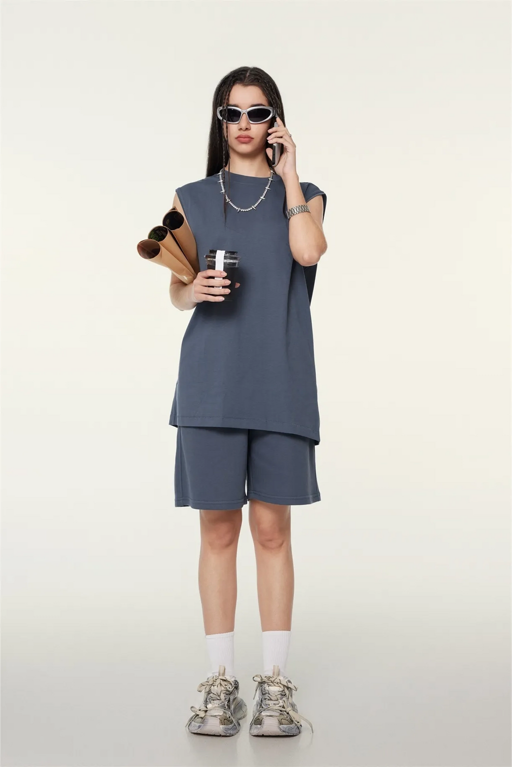 Person wearing a gray sleeveless top and shorts set, holding a coffee cup and sunglasses, on a white background.