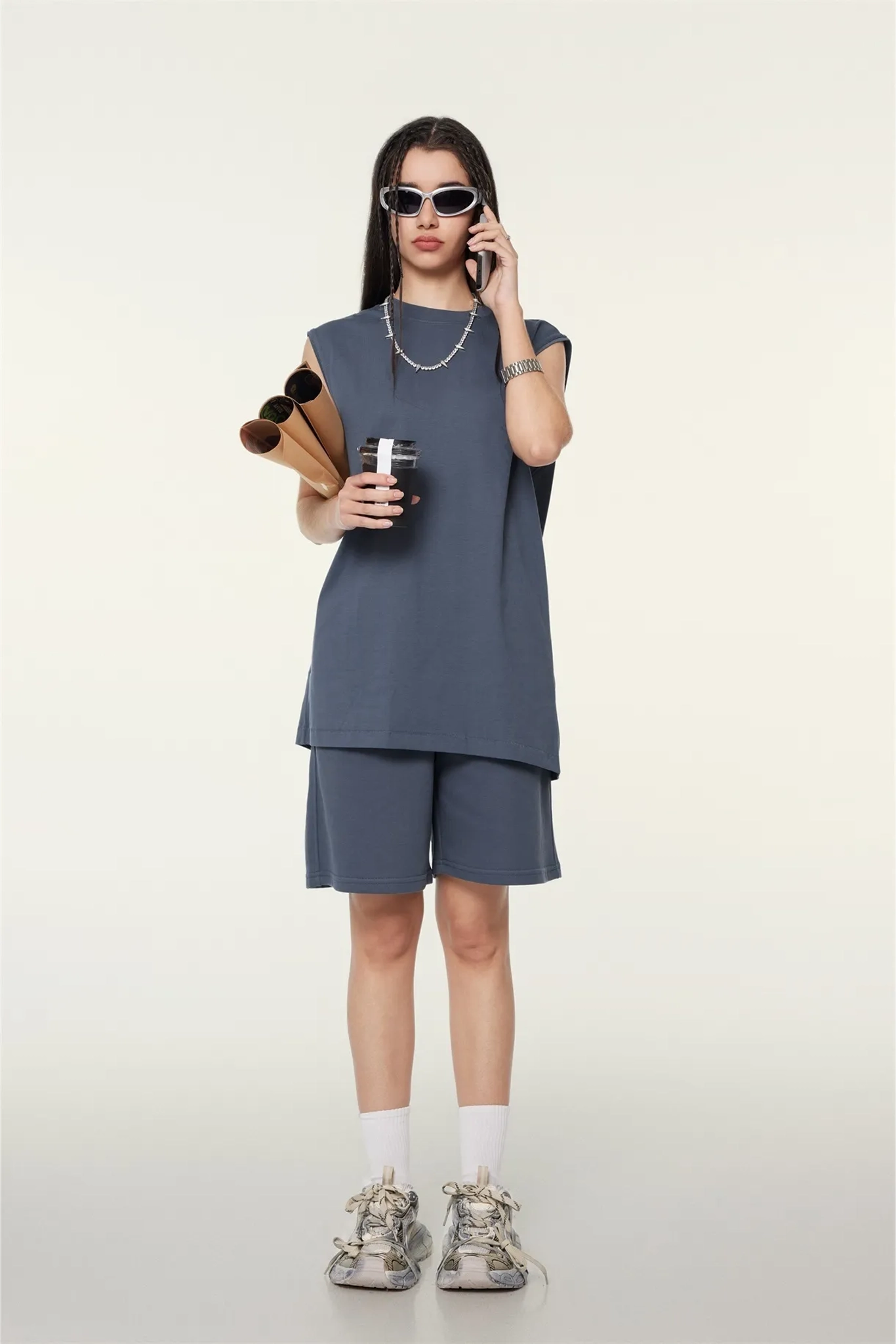 Person wearing a gray sleeveless top and shorts set, holding a coffee cup and sunglasses, on a white background.