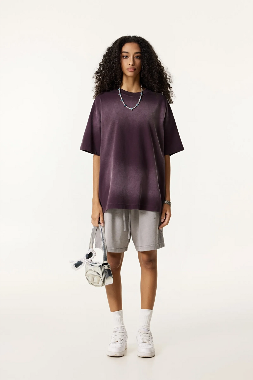 Person wearing a dark purple oversized shirt, light gray shorts, white sneakers, and holding a white bag on a white background.