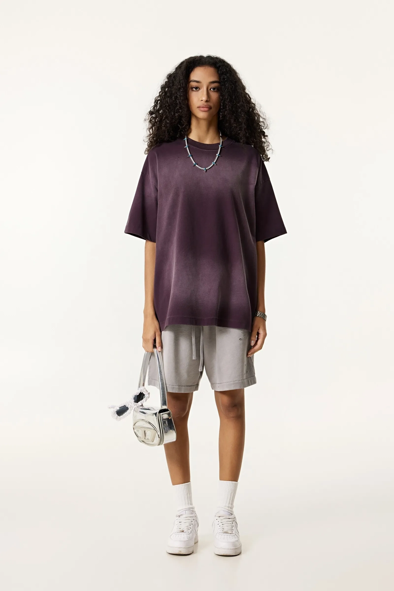 Person wearing a dark purple oversized shirt, light gray shorts, white sneakers, and holding a white bag on a white background.