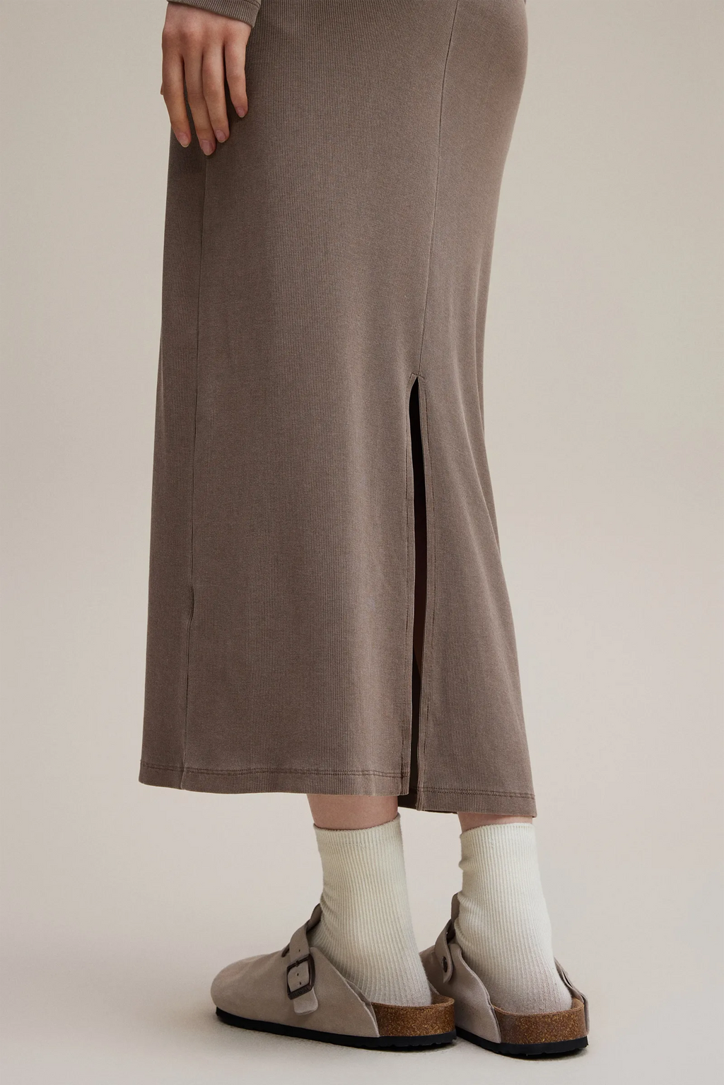 Person wearing a long brown skirt with a slit, white socks, and brown shoes on a beige background